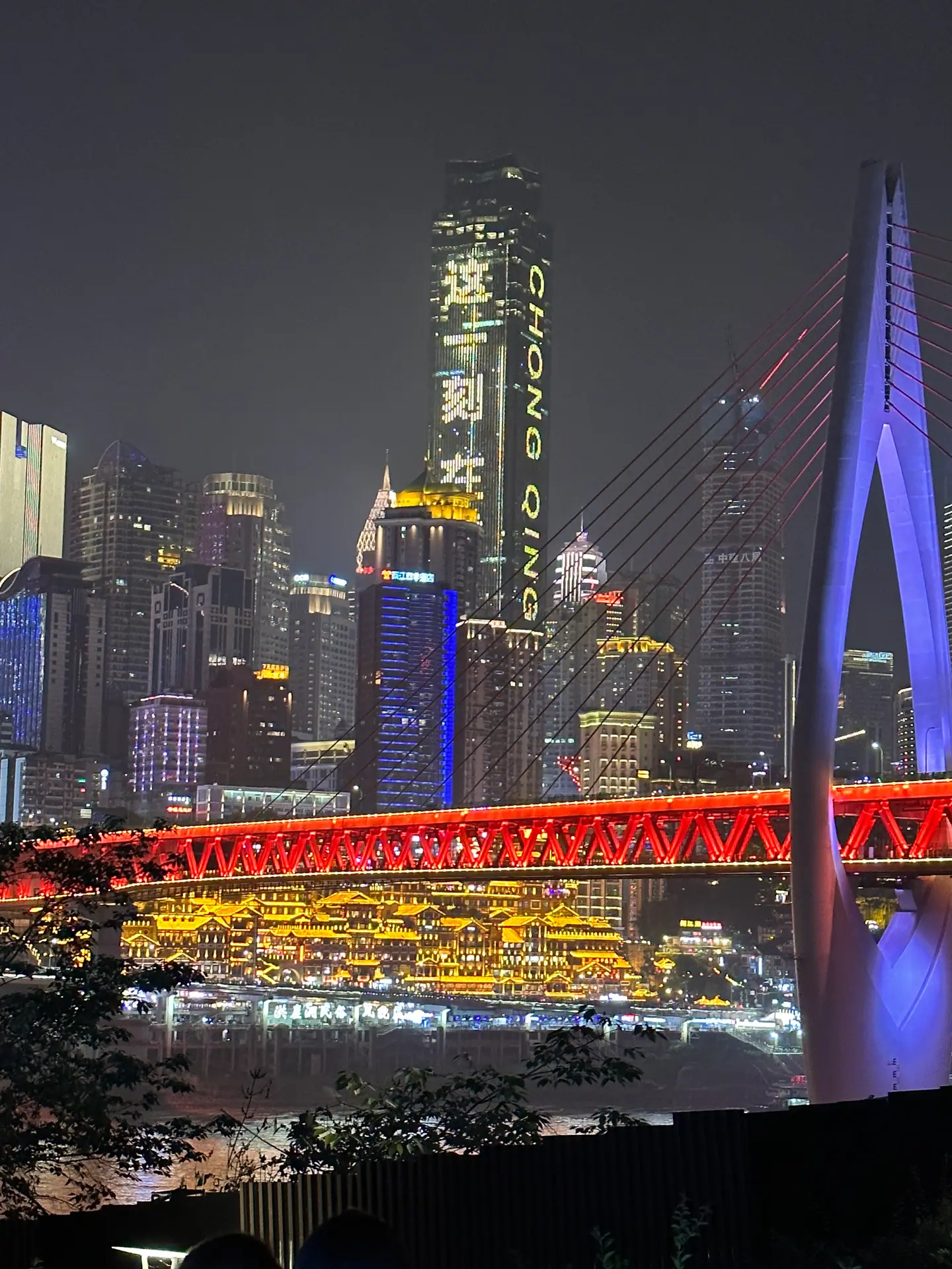 Hongyadong iluminado junto al río bajo un puente rojo, con los rascacielos de Chongqing de noche