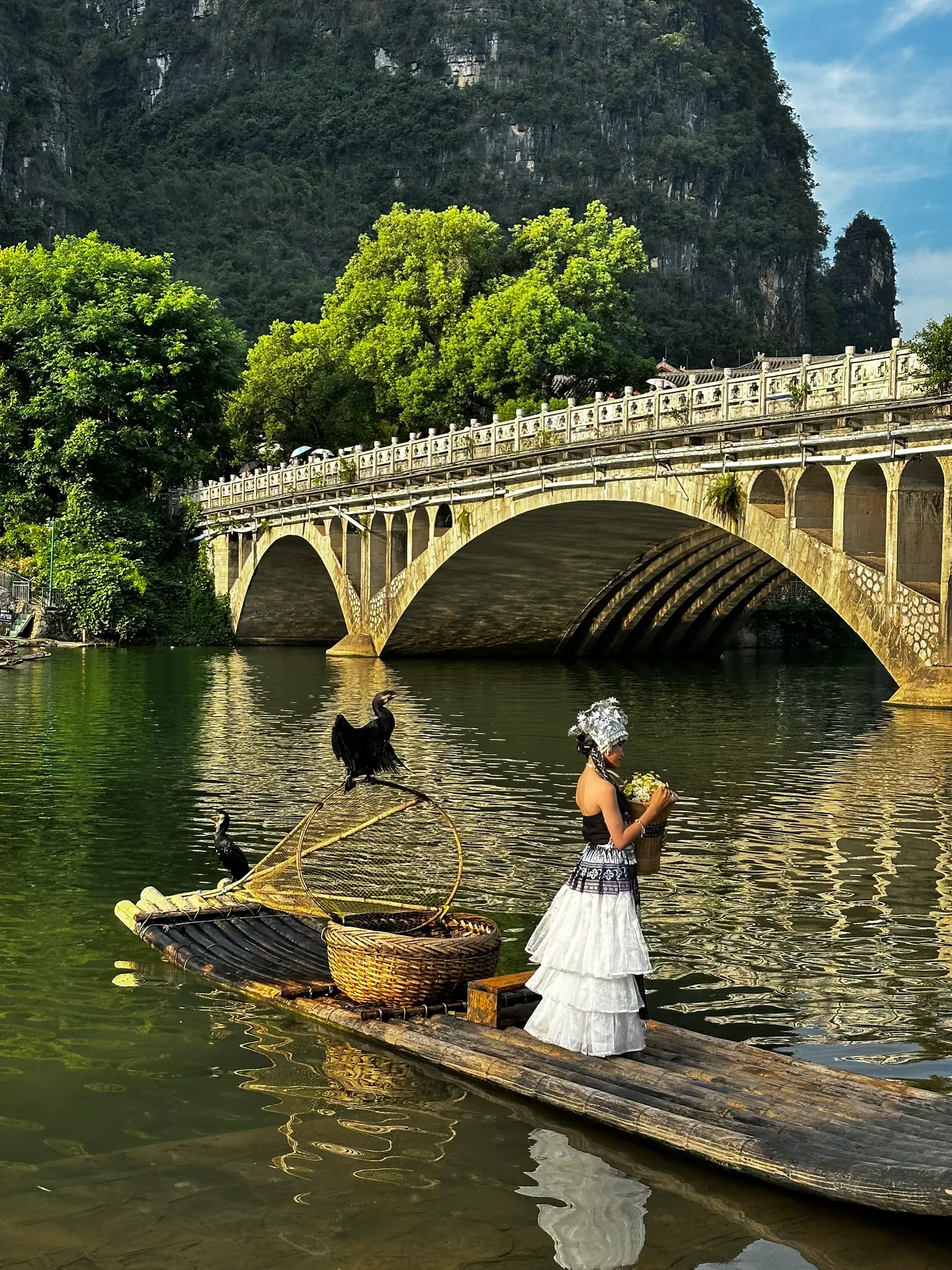 Joven con traje tradicional y cormorán sobre una balsa de bambú bajo un puente de piedra en Yangshuo