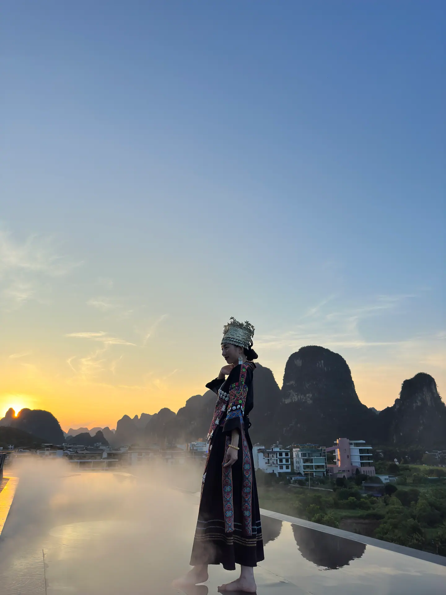 Silueta de una mujer con traje tradicional junto a una piscina infinita frente a los karst al atardecer