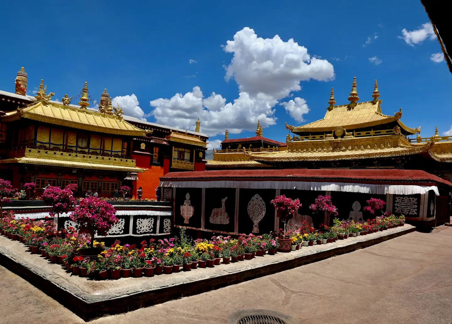 Tejados dorados del templo de Jokhang sobre un patio con macetas de flores en Lhasa, bajo un cielo azul con nubes